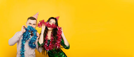Young adult boy and girl 20-25s in party hats pose with New Year's tinsel, make mustaches and look at the camera on a yellow isolated background. Festive banner with copy spaceの写真素材