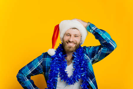Portrait of a young adult guy 20-30s in tinsel, who straightens the santa hat on his head with his hand and looks at the camera with a smile on a yellow isolated background.の写真素材