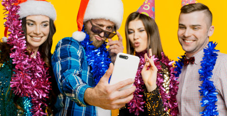 Young adults 20s wearing party hats and santa claus caps and tinsel filming video or taking selfie on smartphone for social media. Selective focus on smartphone in handの写真素材