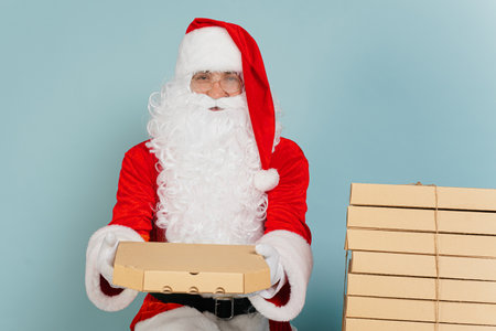 Santa claus holds out a closed pizza box holding it with his hands on a blue isolated background with copy spaceの写真素材