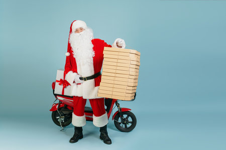 Full length santa claus holds in his hand a huge stack of pizza boxes on the background of a red electro moped on a blue isolated background with copy spaceの写真素材