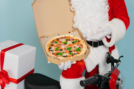 An open box of pizza in the hands of santa claus in white gloves, holds and points to it with his hand against the background of a red scooter on a blue isolated backgroundの写真素材