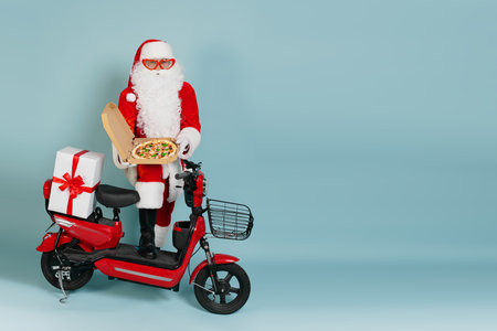 Full length portrait of santa claus in big red heart shaped glasses with an open box of pizza held in his hands in white gloves, against a red moped on a blue isolated backgroundの写真素材
