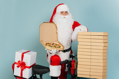 Pizza delivery man dressed as santa claus with an open pizza box in his hand cloaks himself on a stack of boxes on the background of a red scooter and looks at the camera on a blue isolated backgroundの写真素材