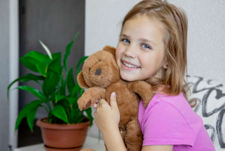Cute little blonde girl with blue eyes hugs a teddy bear, looks at the camera and smiles while sitting on her bed at homeの写真素材