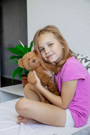A cute little girl gently hugs a plush brown bear, looks at the camera and smiles while sitting on her bed at home.の写真素材