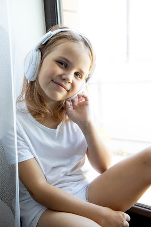 A cute little girl is sitting on the windowsill behind a white curtain at home in her room and...