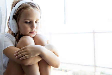 Portrait of a little girl sitting on the windowsill and thoughtfully looking out the window. Hugs his knees and is sad listening to music with headphonesの写真素材