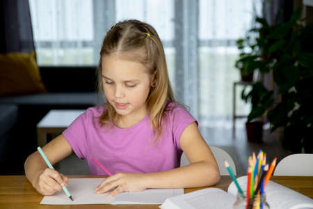 back to school. Cute little blonde girl doing homework in a working school notebook with colored pencils sitting at the table in the living room.の写真素材
