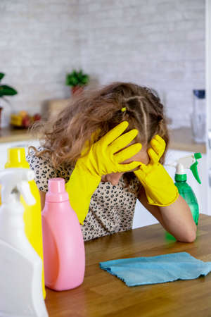 A tired child-a girl with cleaning equipment is sitting at a table in the living room, her head lowered low and her hands on it.The concept of children's housework and cleaning. selective focusの写真素材