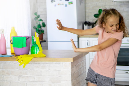 Selective focus on a blonde girl child who shows signs with her hands that she does not want to clean the house. There are detergents, a bucket and rubber gloves on the table.の写真素材