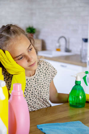 A tired child-a girl with cleaning equipment is sitting at a table in the living room, her head lowered low and her hands on it.The concept of children's housework and cleaning. selective focus.の写真素材