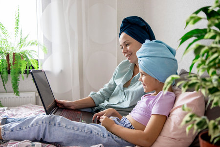 A beautiful young woman and her daughter are using a laptop at home, lying on a bed with towels on their heads.の写真素材