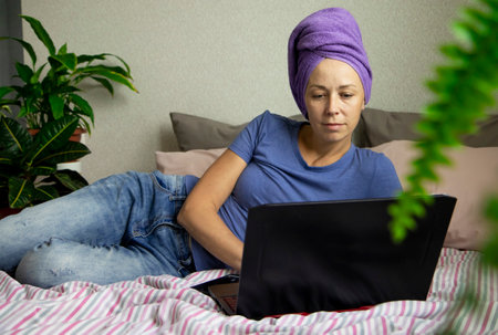 a young woman is working on a laptop at home, lying on the bed in her bedroom with a towel on her head. The concept of working from home, remote online work via the Internetの写真素材