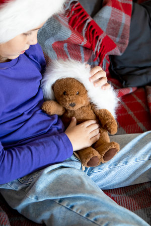 A cute little girl in a Santa hat is playing and hugging a teddy bear while sitting on the sofa in the living room. The concept of family holidays and traditions, love and careの写真素材