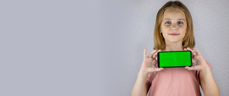 Portrait of a little blonde haired girl showing a mobile phone application in her hands with a green screen. Banner, place for text, copy space.の写真素材