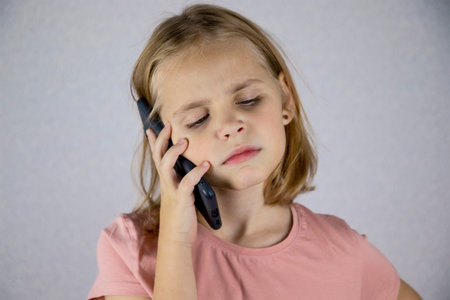 A cute little girl calls on a mobile phone, has an unpleasant conversation. Bad emotions of a child when communicating on a smartphoneの写真素材
