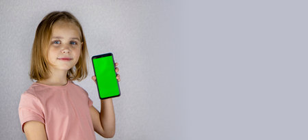 Portrait of a little blonde haired girl showing a mobile phone application in her hands with a green screen. Banner, place for text, copy space.の写真素材