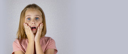 Frightened little girl looks at the camera worried about something with her hands on her face and her eyes wide open. banner, copy spaceの写真素材
