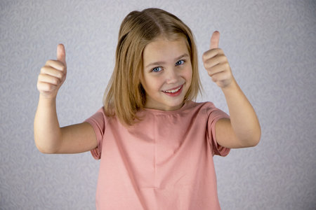 Portrait of a laughing little girl with blond hair showing a thumbs up with both hands. Hand signs and gesturesの写真素材