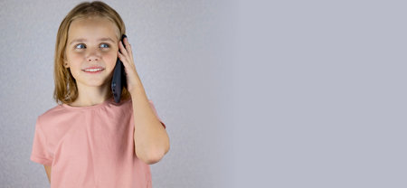 A cute little girl calls on a mobile phone, listens and maintains a conversation. Vivid emotions of a child when communicating on a smartphone. Banner.の写真素材