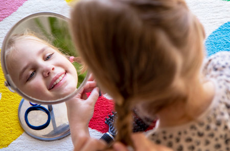 The older sister braids the braids of the younger sister. A little girl looks in the mirror at her reflection. Hair care, hairstyles for childrenの写真素材