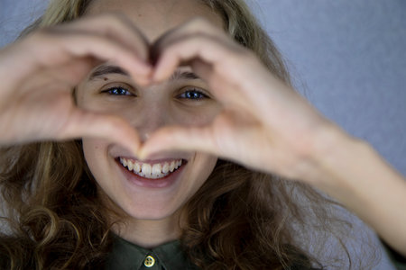 A cute teenage girl makes a heart shape with her hands and fingers and smiles joyfully. Good mood and expression of loveの写真素材