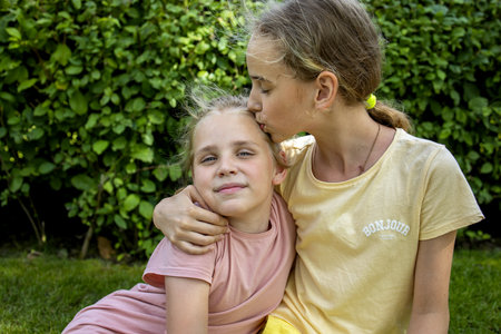 Two girls-sisters of school age hug each other. The older one kisses the younger one. A walk through the summer city park. Love and friendship in the familyの写真素材