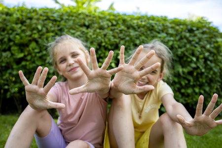 Two girls-sisters of school age are sitting on a green lawn while walking through a summer city park and showing their palms. Love and friendship in the family.の写真素材