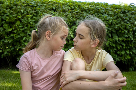 Two girls-sisters of school age are sitting on a green lawn while walking through a summer city park and chatting. Love and friendship in the family.の写真素材