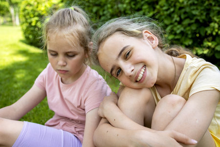 Two girls-sisters of school age are sitting on a green lawn while walking through a summer city park and chatting. Love and friendship in the family.の写真素材