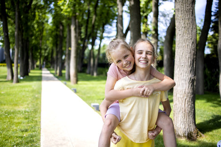Two girls sisters of school age hug each other. They laugh and fool around. A walk through the summer city park. Love and friendship within the family.の写真素材