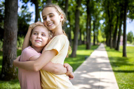 Two girls sisters of school age hug each other. A walk through the summer city park. Love and friendship within the familyの写真素材