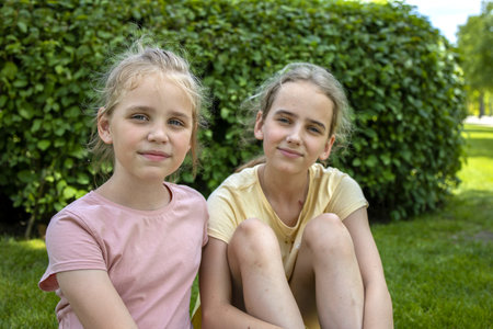 Two girls-sisters of school age are sitting on a green lawn while walking through a summer city park and chatting. Love and friendship in the family.の写真素材