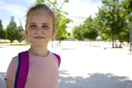 Portrait of a little blonde girl with a backpack. A young tourist walks through the summer park in a good mood. The concept of walking and a healthy lifestyle.の写真素材