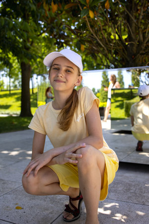 Portrait of a pretty blonde teenage girl in a cap on a background of mirrors. A walk through a beautiful summer park. Beautiful smile and good mood.の写真素材