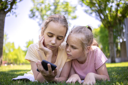 Two cute blonde little girls are relaxing in the park lying on their stomach. They watch videos or social networks, send messages using a smartphone. Available wireless internetの写真素材