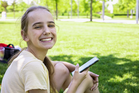 A beautiful blonde teenage girl is resting in a city park on the lawn, watching videos or social networks, sending messages using a smartphone. Affordable wireless internet for teenagers.の写真素材