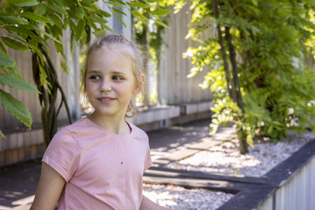 portrait of a cute little girl against the background of green plants on the street. The child looks away and smiles. place for text.の写真素材