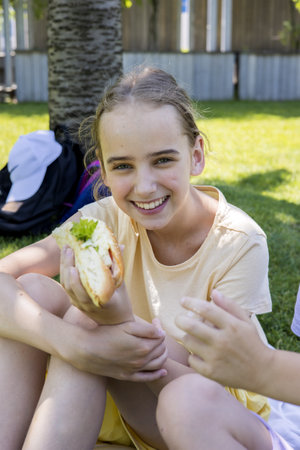 A teenage girl with blonde hair is sitting on the lawn in a summer park and eating a delicious and nutritious sandwich. The concept of urban foodの写真素材