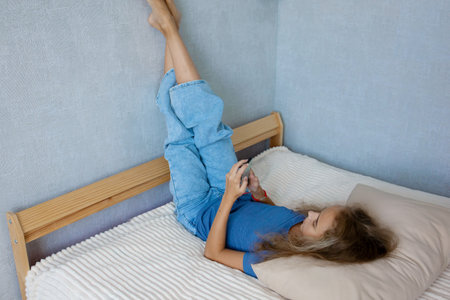 Cute teen girl browsing social media while lying on her bed in the bedroom. Communication of modern childrenの写真素材