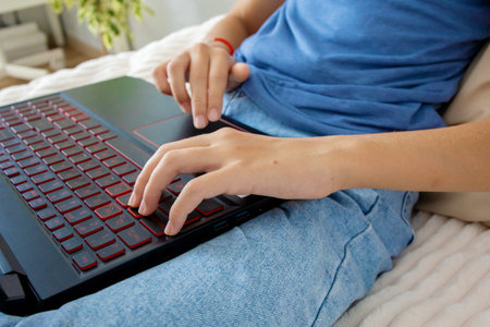 Cropped photo of a teenage girl typing text on a laptop while sitting at home on her bed.