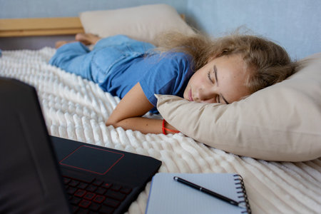 Tired of lessons, a blonde teenage girl sleeps on the bed next to her laptop. The concept of home education and self-studyの写真素材