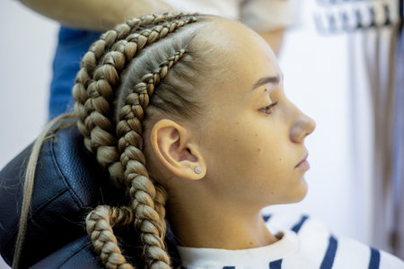 A girl sits in a modern hair salon chair, her hair intricately braided. The soft light highlights her focused expression while a stylist works expertly nearby.の写真素材