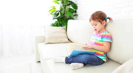 cute baby girl reading a book at home sitting on the sofaの写真素材