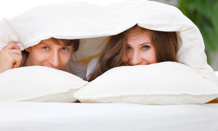 Happy couple laughing in bed peeking out from under the blanketの写真素材