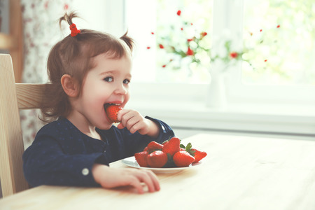 happy child girl eats strawberries in the summer home kitchenの写真素材
