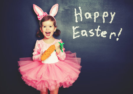 Happy Easter! child girl in costume bunny rabbit with e carrot about blackboard