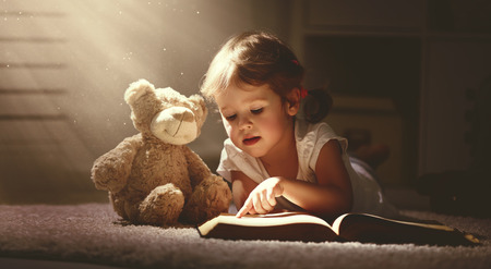 child little girl reading a magic book in the dark home with a toy teddy bearの写真素材
