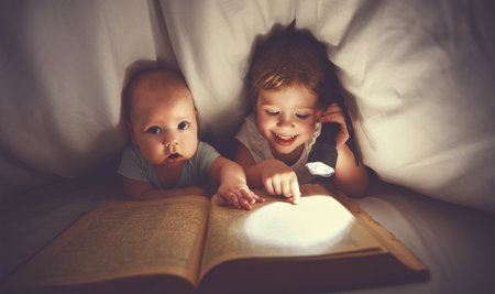 children brother and sister read a book with aflashlight under blanket in bedの写真素材